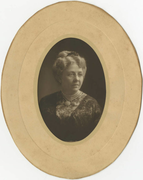 Black and White photograph of a white woman in her 50s or 60s with her hair in a bun, wearing peals and wire-framed glasses. The photo is inset in an oval beige frame.