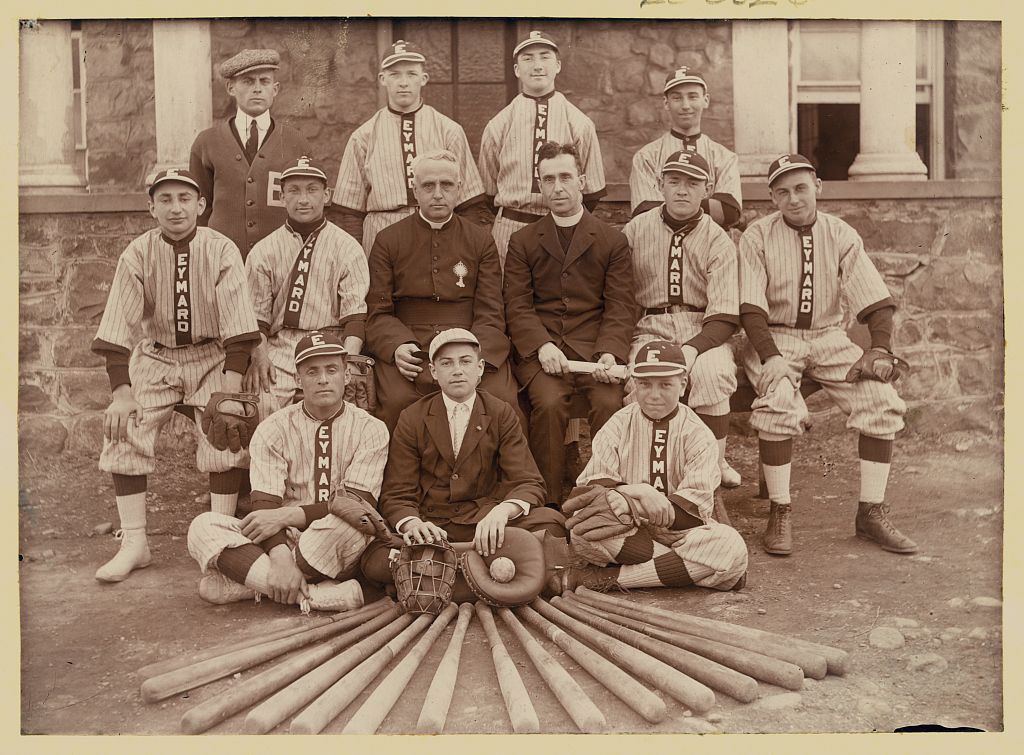 Photograph shows the Eymard Seminary baseball team wearing uniforms, gathered around two clergymen, one holding a biretta, all posed in front of a stone building with a display of baseball bats, a catcher's mask, and a baseball in the foreground.