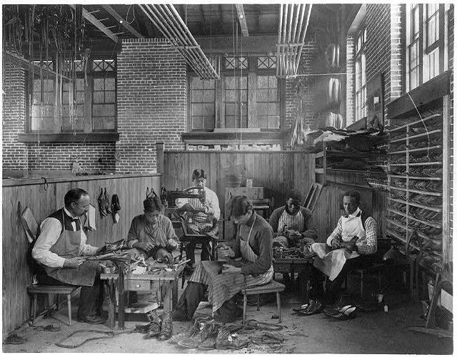Black and white photograph of five men making and repairing shoes in a workshop that has a wall of shoes.