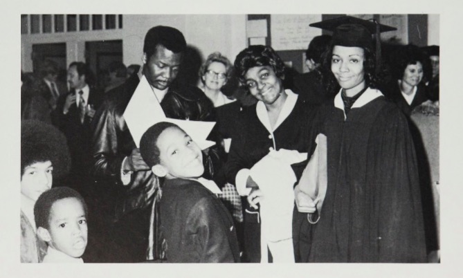 A Black female student smiles at the camera in graduation attire, with her family grouped and smiling to her right.