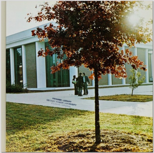 A young red-leafed tree with sunlight filtering through the leaves stands before a white and brick building with large glass windows. Three people talk near the building in bell-bottomed pants.