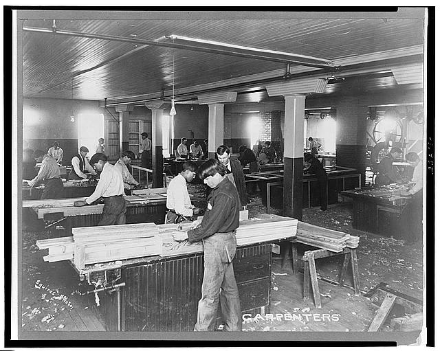 Black and white photograph of many students standing at a variety of workstations working with wood. Some teachers are walking around to help students.