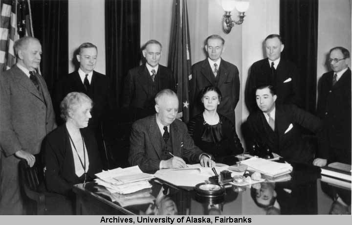 Black and white photograph of Governor Troy writing on a piece of paper. He is sitting at a table with two people on his left, and one woman on his right. Six men are standing behind him, and all are wearing formal attire.