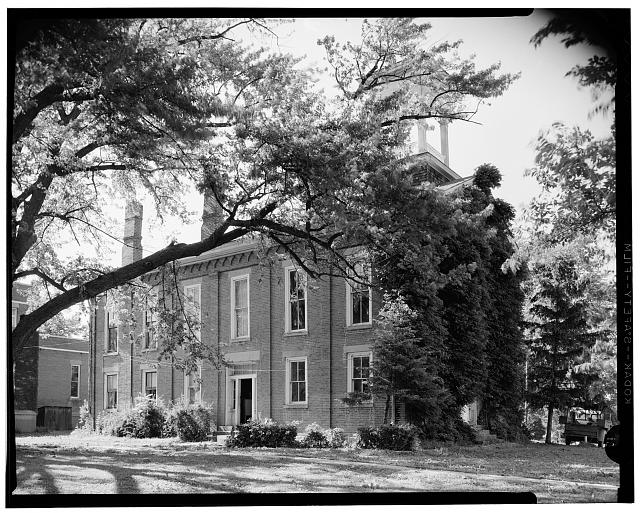 Black and white photograph of a two story building which is framed by a tall tree in the photograph.