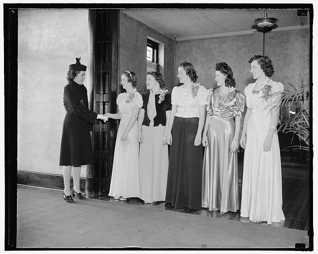 Black and white photograph of 5 women standing in a line, wearing very formal dresses. Another woman is walking in front of them and shaking the first women's hand.