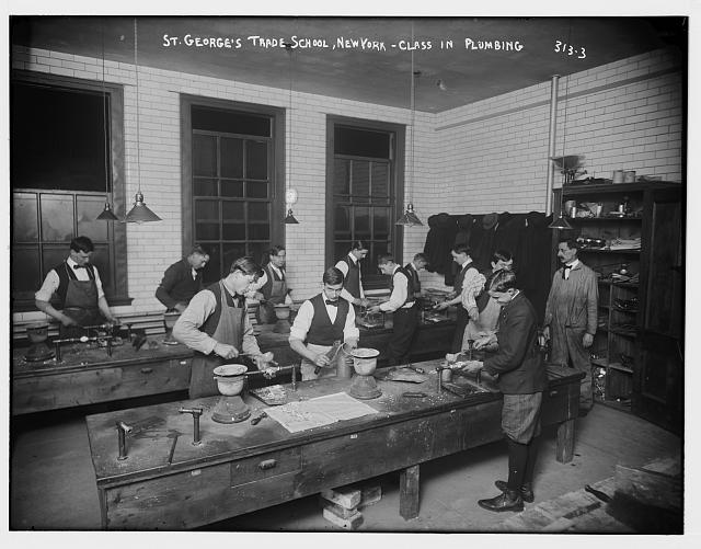 Black and white photograph of a classroom where several students are standing at stations and working with various pieces of plumbing.