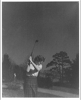 Black and white photograph of a woman who is swinging a golf club in the air, while on a golf course.