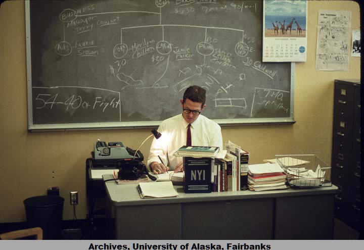 Color photograph of an University of Alaska employee sitting at a crowded wooden desk in front of a chalkboard, working on some papers.
