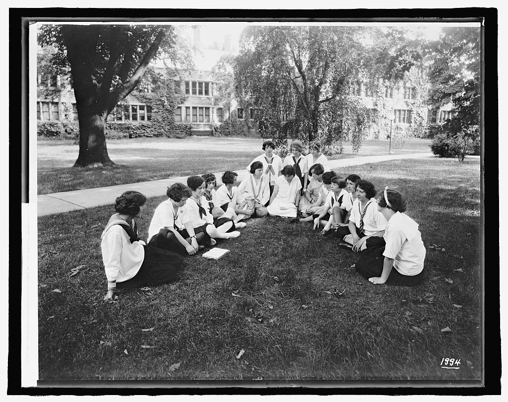 Group of women wearing uniforms sitting in a v-shape on a grassy lawn. Behind them are buildings and trees of the campus.