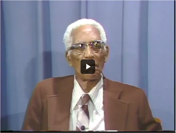 Screenshot of a video showing an older Black man with white hair and glasses wearing a brown suit being interviewed.